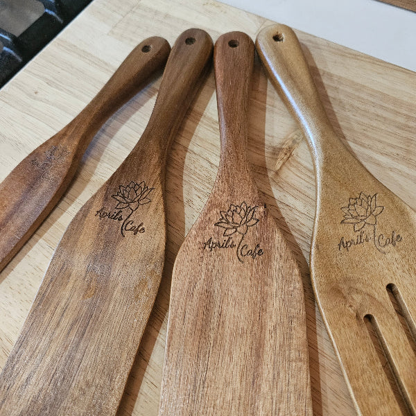 A set of four engraved wooden chef's spatulas of different sizes, displayed on a wooden surface. Each spatula has a handle with a personalized engraving of a flower and the text 'APRIL CAFI'.